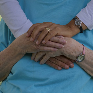 Caregiver tiene le mani di una persona anziana seduta, immagine legata all’assistenza e alla longevità.