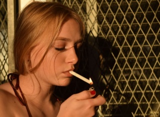 Giovane donna di profilo accende una sigaretta con un accendino davanti a una grata metallica, illuminata da luce calda.